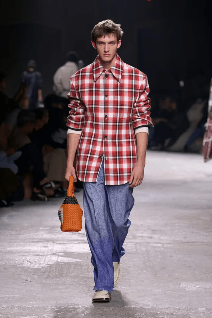A male model walks the bottega venetta 25ss runway wearing a red and white checked oversized shirt, paired with relaxed denim pants and holding an orange woven bag.