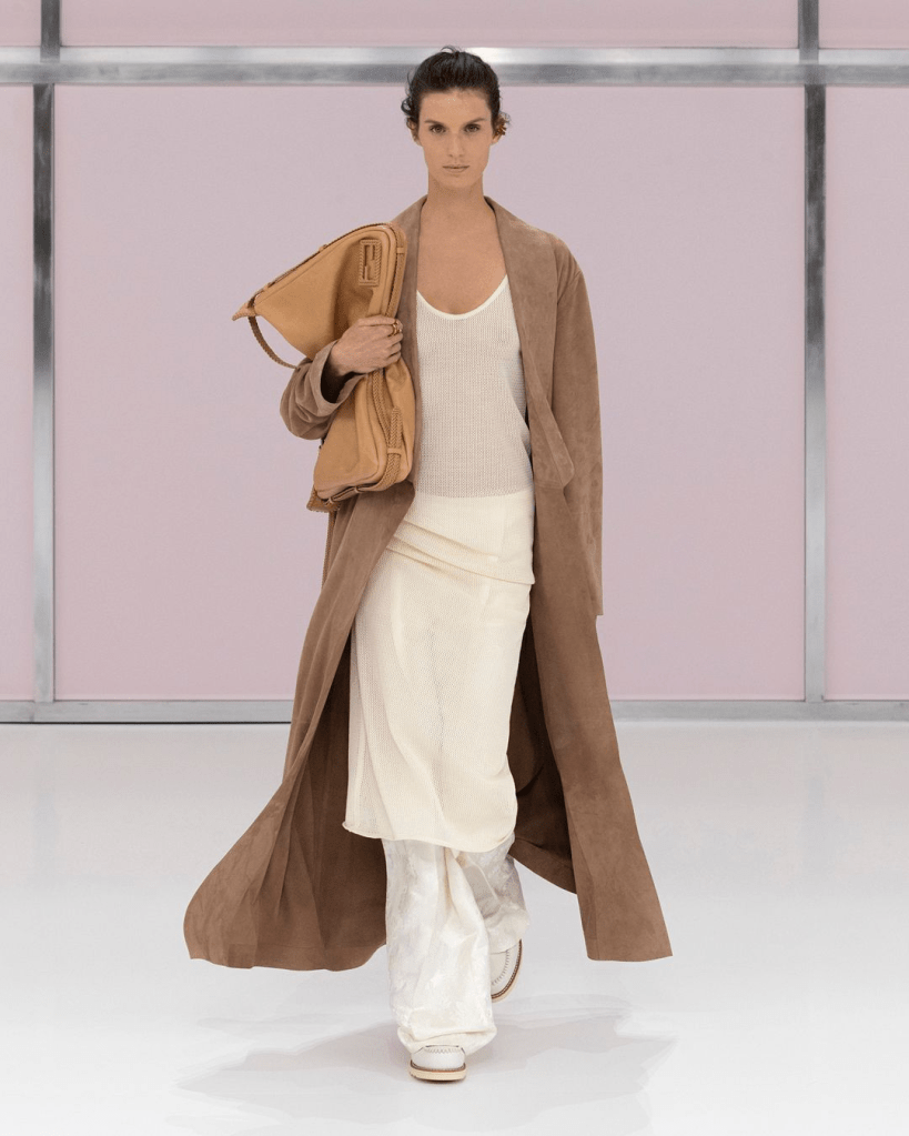 A model walking down Fendi 25SS runway wearing a long brown coat, a white top, and light-colored pants, holding a large brown handbag, with a pastel pink background.