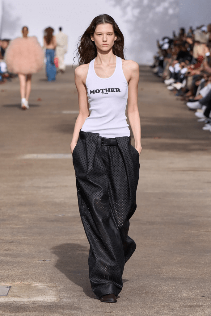 A model walks down the Stella Maccartney 25ss runway wearing a white tank top with 'MOTHER' printed on it and loose, black, perforated trousers during a fashion show.