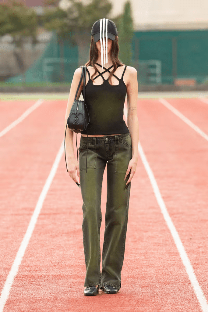 A model poses on AVAVAV 25SS runway, wearing a black tank top with unique strap detailing, olive green straight-leg jeans, a baseball cap with white stripes, and carrying a small black handbag.