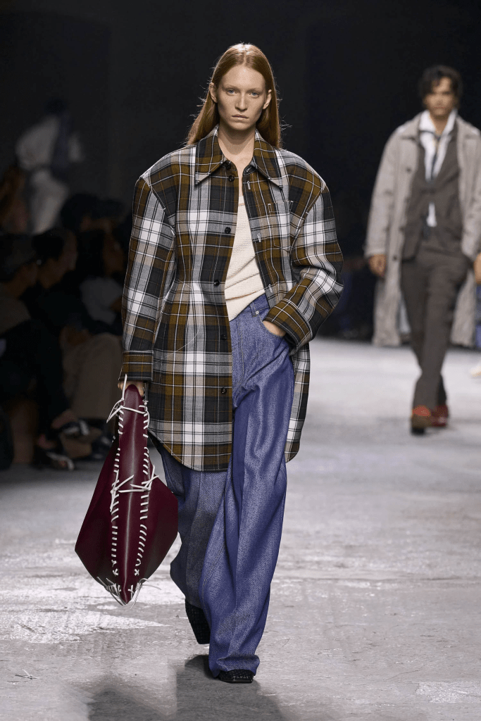 Model walking on the Bottega Venetta SS25 runway wearing a plaid jacket, light top, and wide-leg trousers, carrying a large bag.