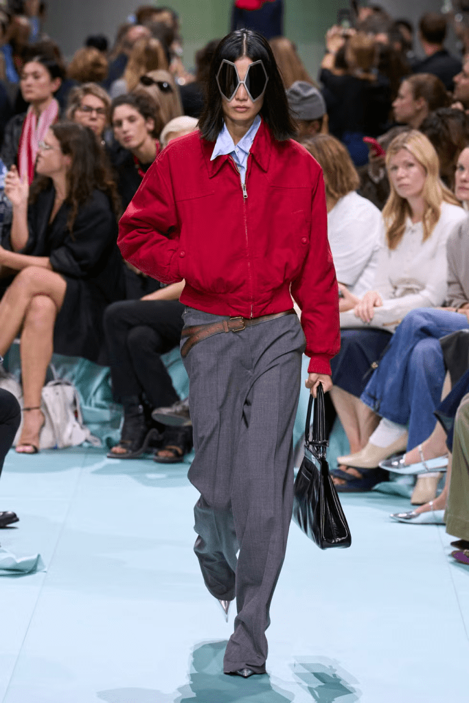 A model walks down a Prada 25SS runway wearing a red bomber jacket, gray trousers, and oversized sunglasses, showcasing a blend of urban streetwear and high fashion, with an audience of spectators in the background.
