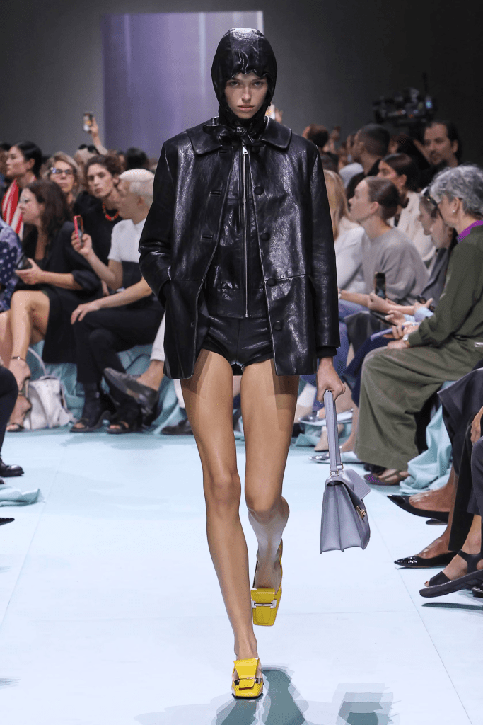 A model walks down the Prada 25SS collection runway wearing a shiny black outerwear piece with a hood, paired with shorts and bright yellow shoes, while an audience observes.