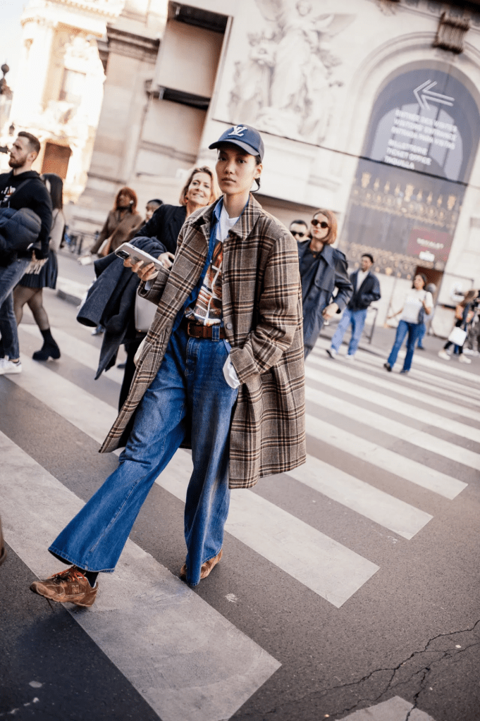 A stylish individual walks on a city crosswalk, wearing a plaid coat and vintage denim jeans, holding a smartphone while reflecting modern fashion trends.
