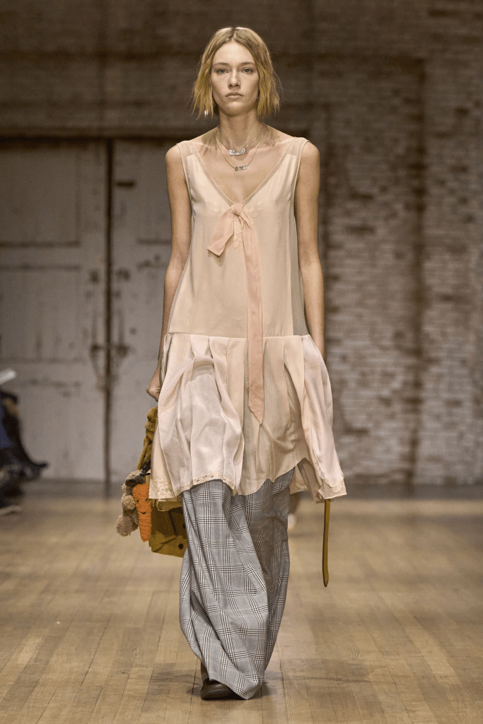 A model walking on the Coach 25fw runway wearing a light pink sleeveless dress with a bow detail and loose plaid trousers, showcasing a blend of elegance and casual style.