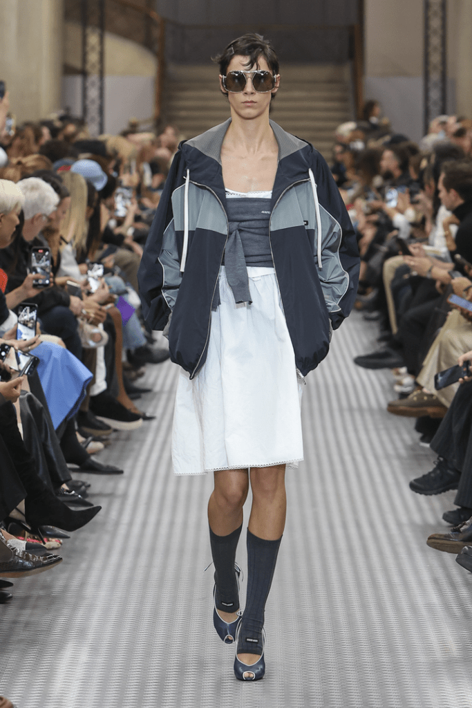 A model walks down the miumiu 25ss runway showcasing a contemporary athleisure outfit, featuring a stylish oversized track jacket, fitted top, and a flowing skirt, while wearing sunglasses and fashionable footwear.