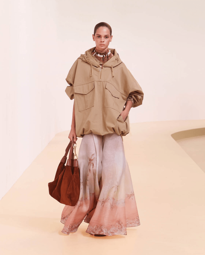 A model walks down the Zimmermann 25SS runway wearing a tan oversized hooded sport jacket paired with a flowing, pastel-colored maxi skirt and carrying a brown bag.