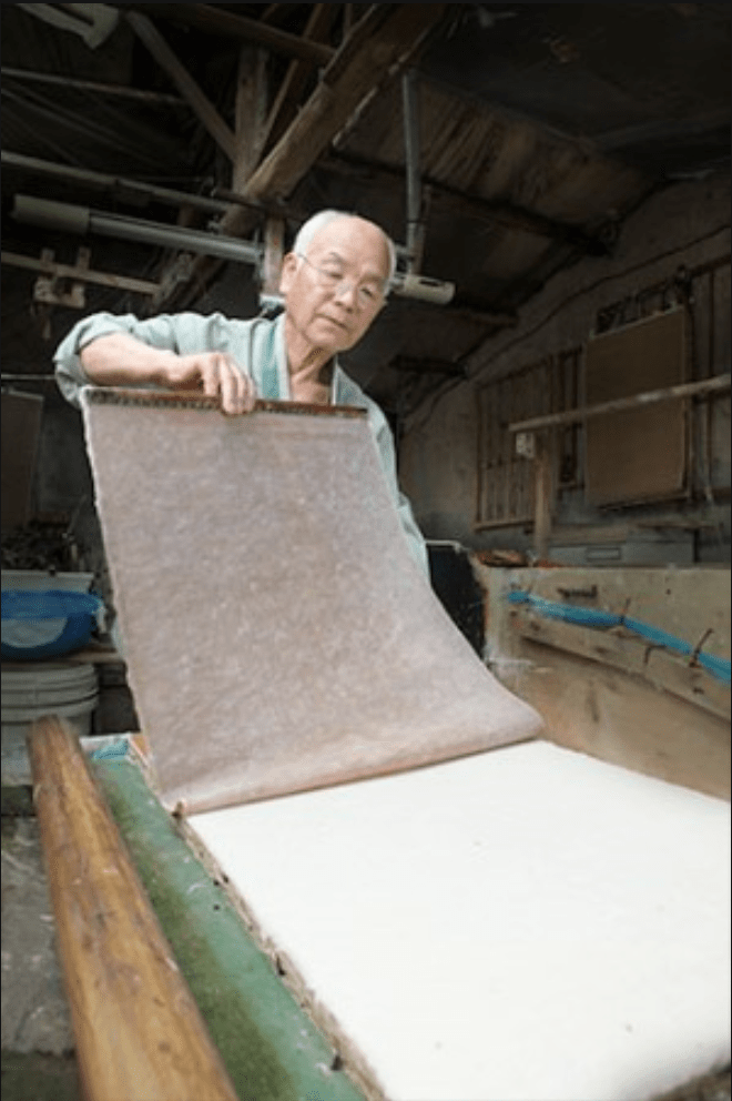 An artisan carefully holds a sheet of traditional Korean hanji paper in a workshop, showcasing the craftsmanship behind this centuries-old material.