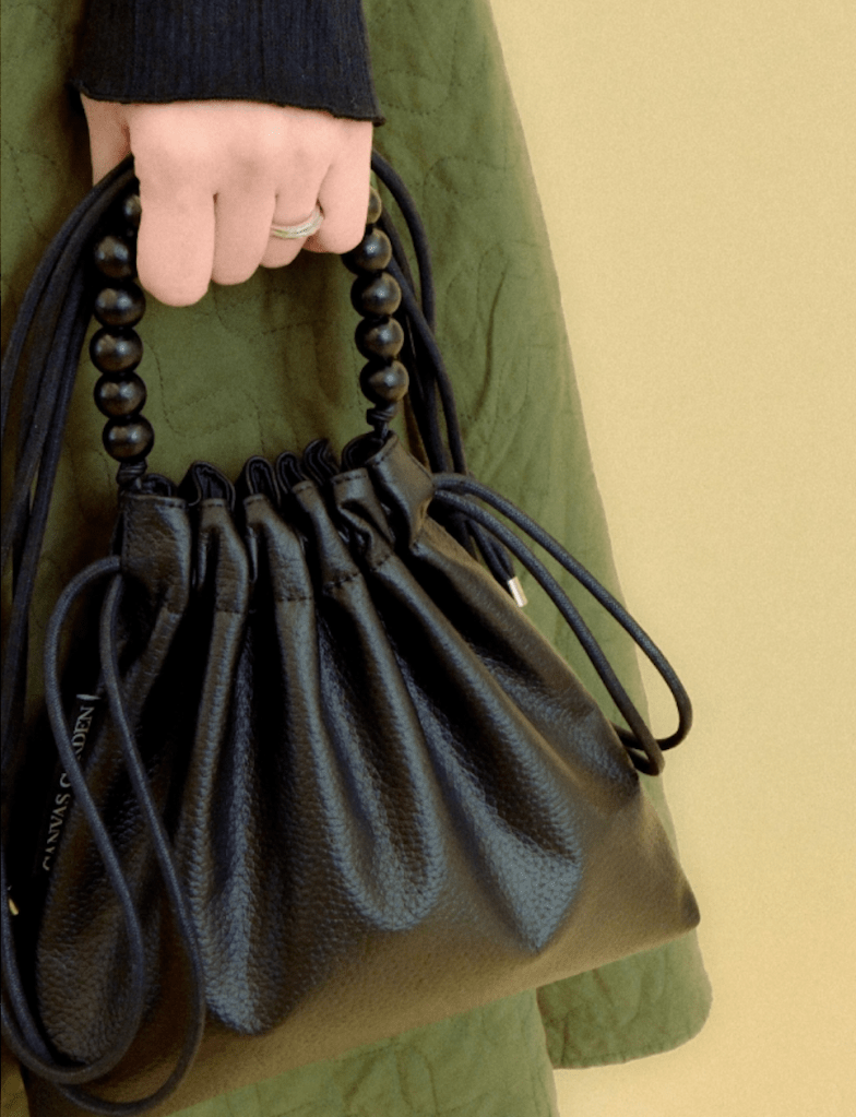 A close-up of a hand holding a black handbag with a gathered design and beaded handle, set against a green quilted background.