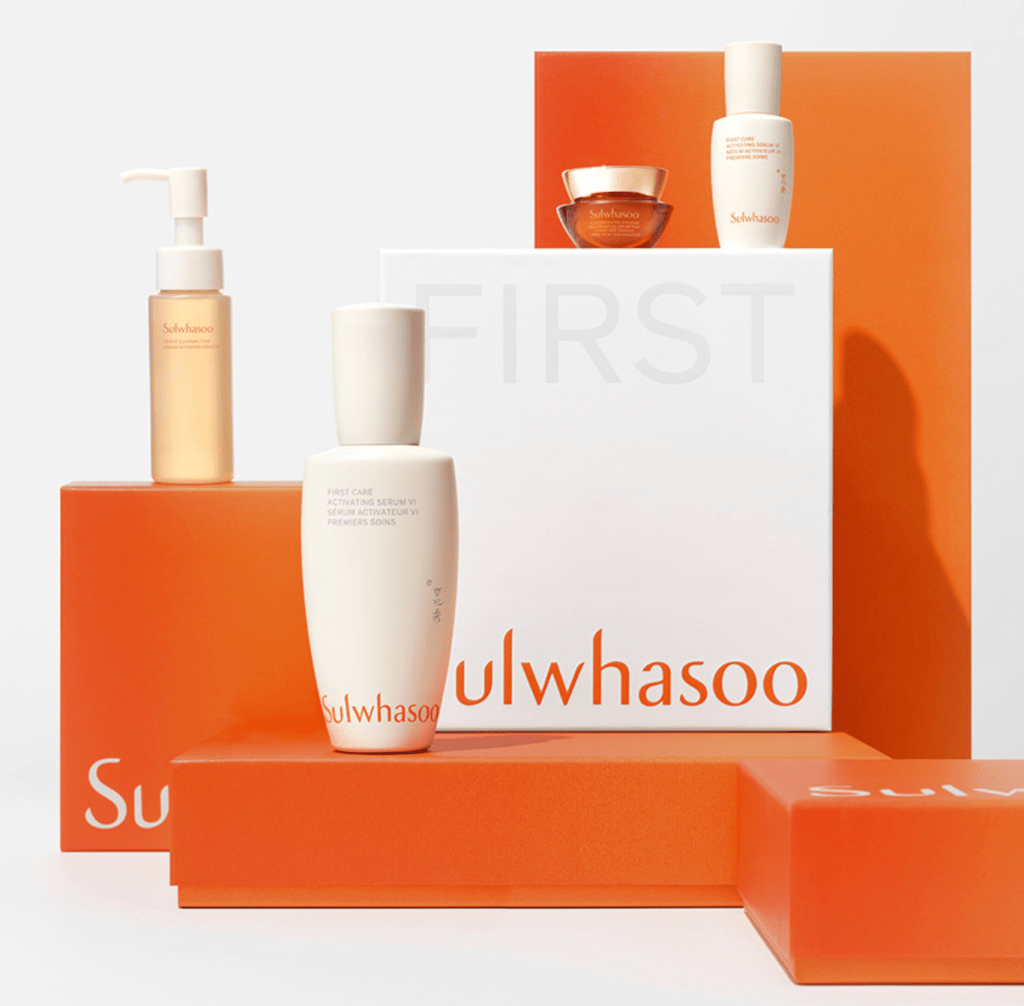 A collection of skincare products from the brand Sulwhasoo, featuring a white bottle labeled 'FIRST CARE ACTIVATING SERUM', a small golden jar, and other assorted bottles, all presented against an orange gradient background.