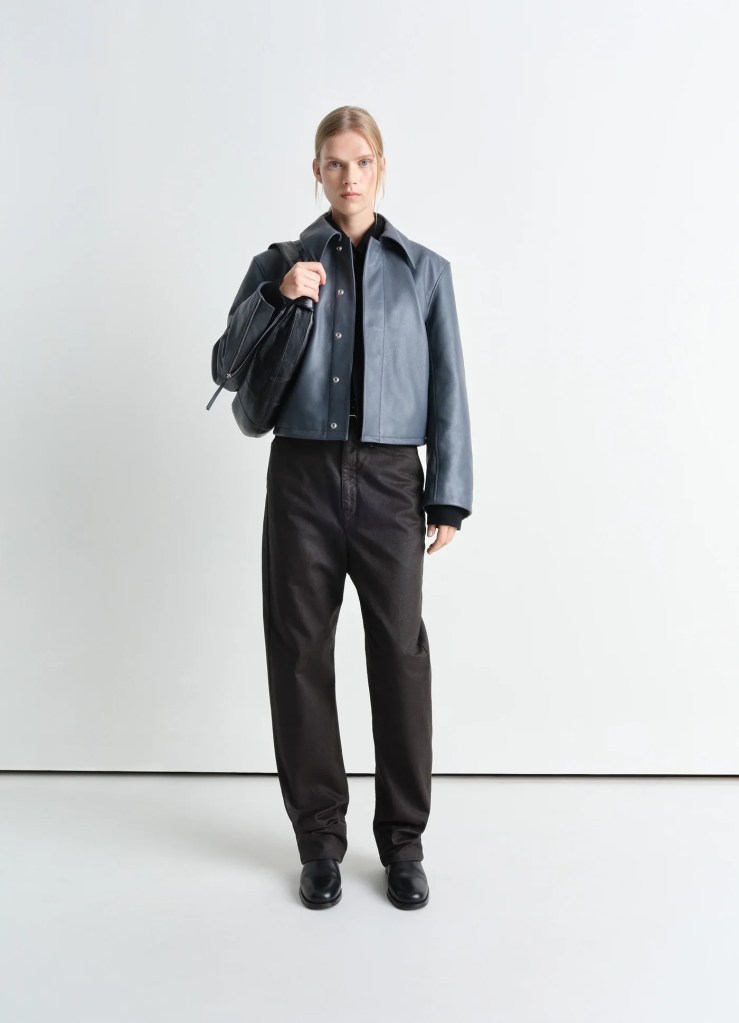 A model standing against a plain background, wearing Lemaire 25SS collection a cropped leather jacket with a collared shirt and black trousers, holding a handbag.