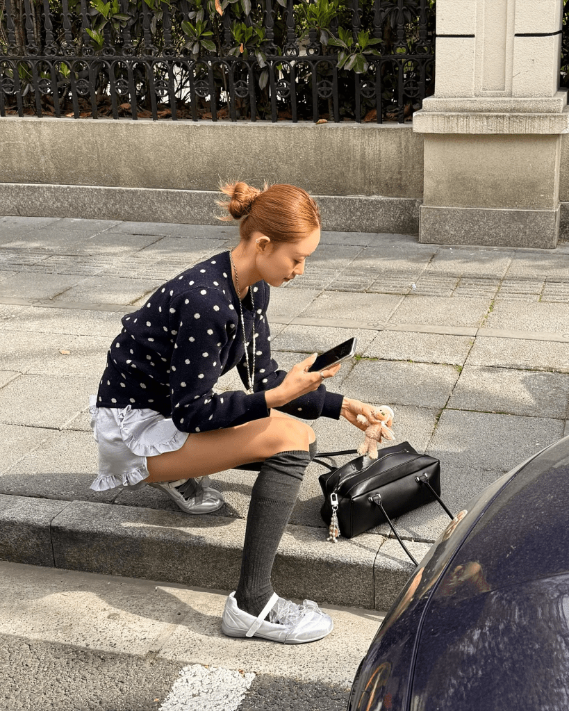 A young woman kneels on the sidewalk, wearing a polka dot sweater, ruffled skirt, and knee-high socks paired with ballet flats. She is focused on her phone while holding a small toy and has a black handbag beside her.