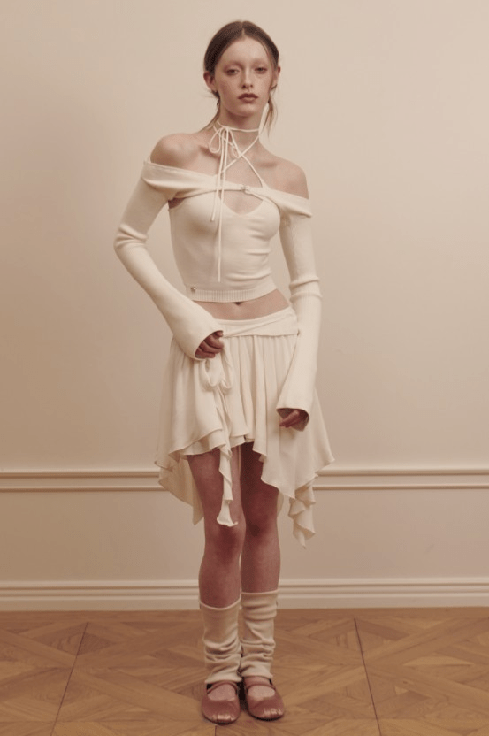 A model wears a soft cream off-shoulder top paired with a flowing skirt and leg warmers, embodying the balletcore aesthetic.