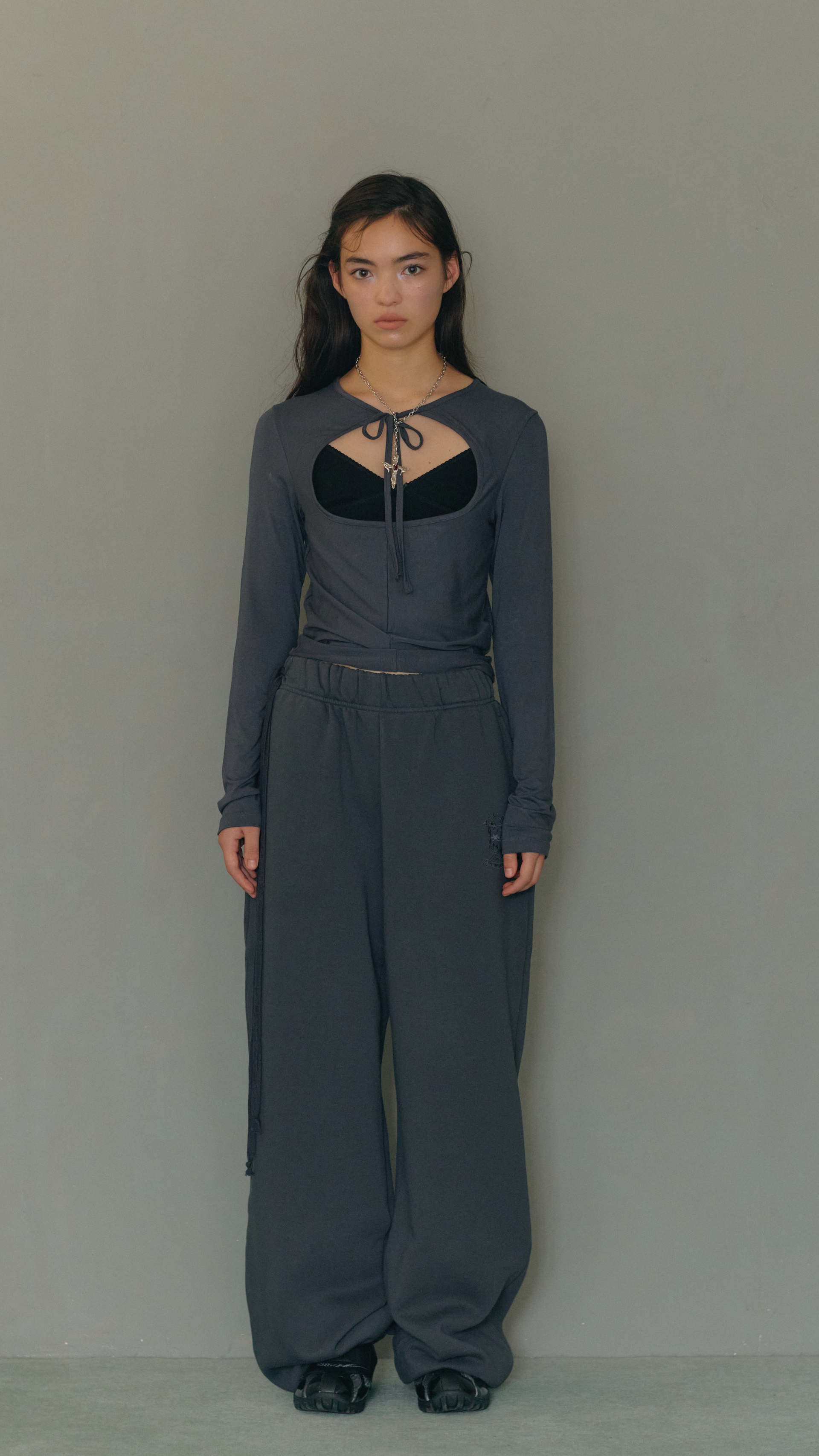 Young woman wearing balletcore style look a gray long-sleeve top with cut-out details and wide-leg pants, posing against a plain background.
