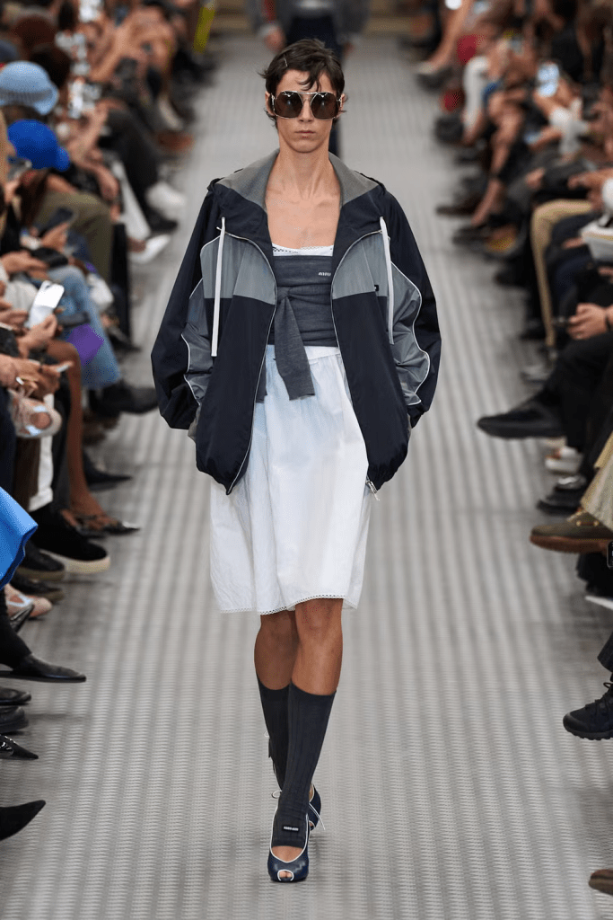 Model walking down a Miumiu 25SS collection runway wearing a stylish, oversized track jacket paired with a white dress and knee-high socks, surrounded by an audience.