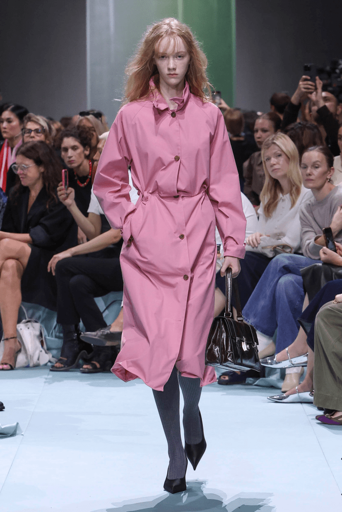 Model showcasing Prada 25SS collection a stylish pink trench coat during a fashion show, with a modern twist and minimalist accessories.