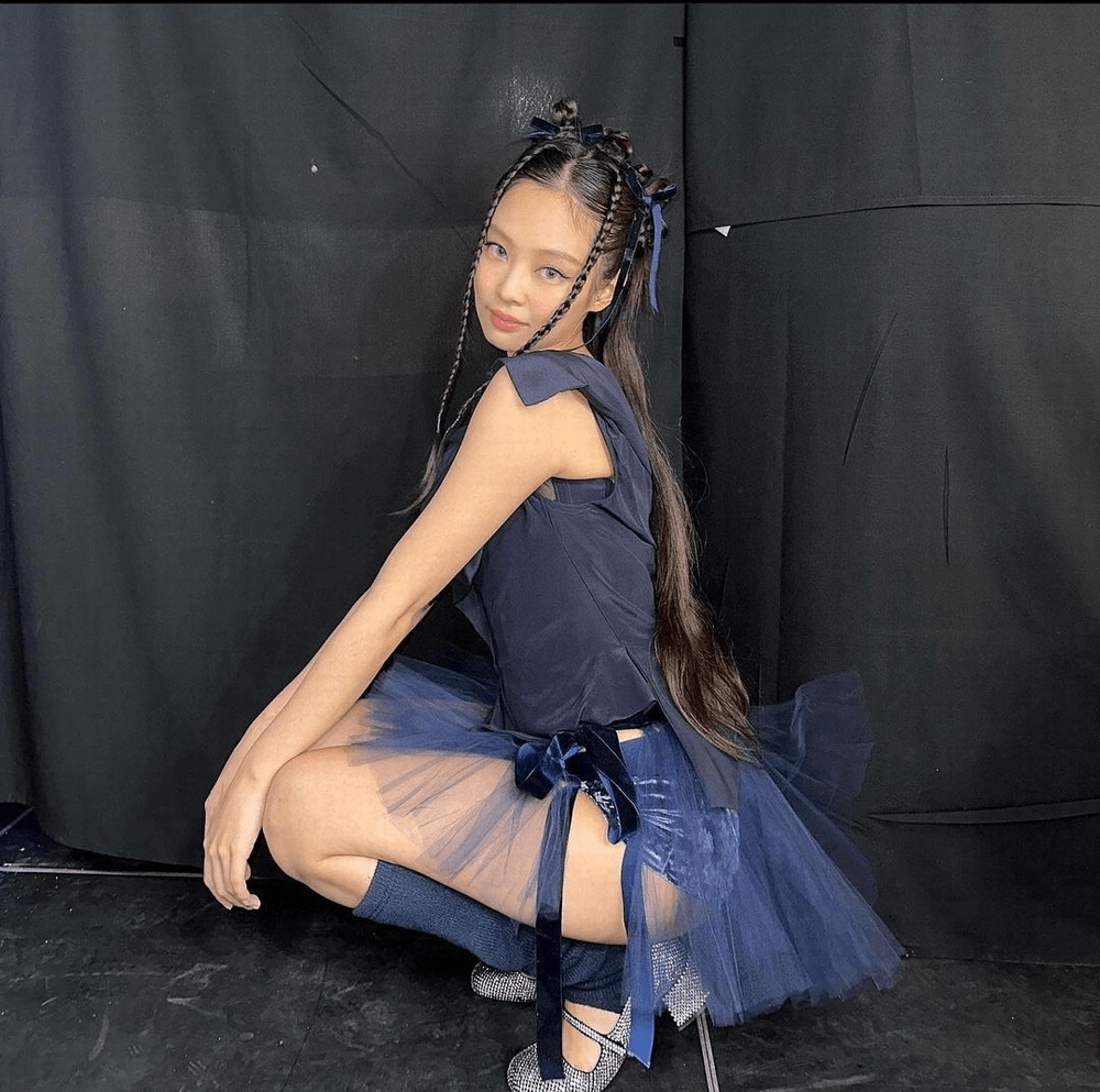 Jennie wearing a navy ballet outfit with a layered tulle skirt, crouching gracefully. Her long hair is styled with braids and ribbons, complemented by thigh-high socks and embellished shoes.