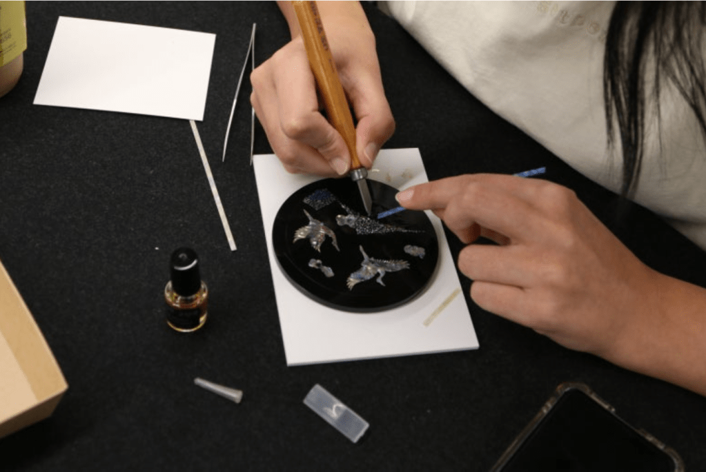 A person carefully applying mother-of-pearl inlay to a black lacquer piece, surrounded by various tools and materials involved in the jagae craft process.