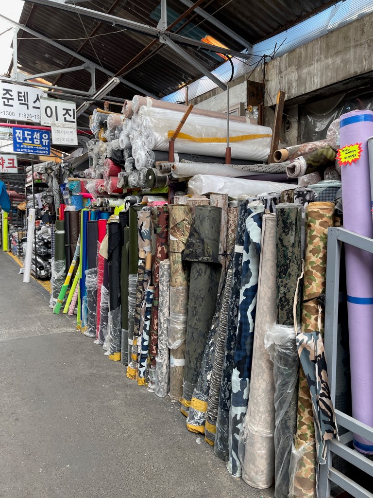 Stacks of various fabric rolls displayed in an outdoor market, showcasing colorful textiles in sinseol-dong material market.