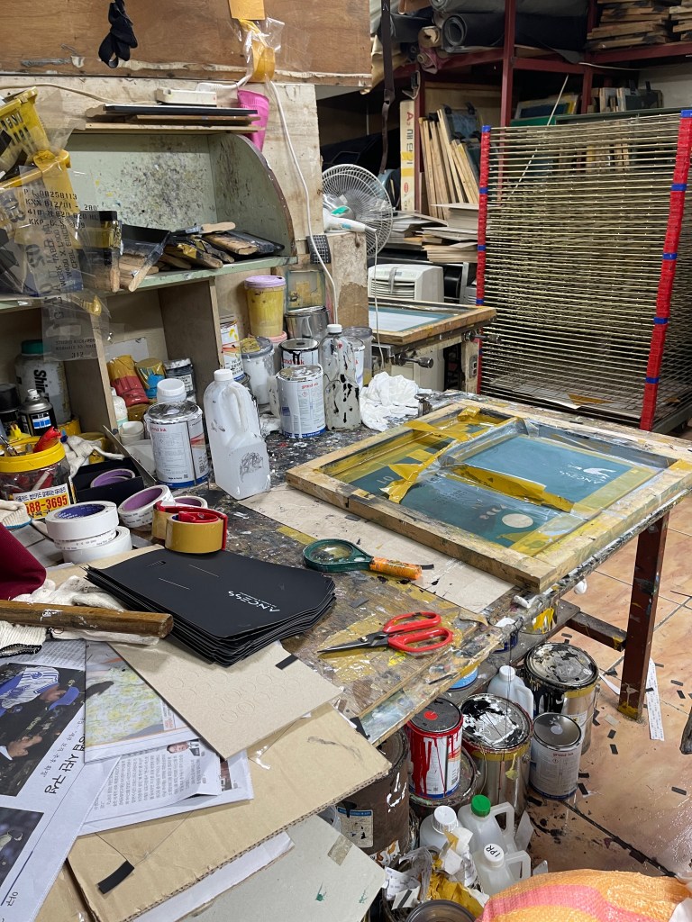 A cluttered stamping workspace featuring paint cans, brushes, scissors, and a wooden board with vibrant colors, showcasing the creative chaos of textile design in sinseol-dong material market.