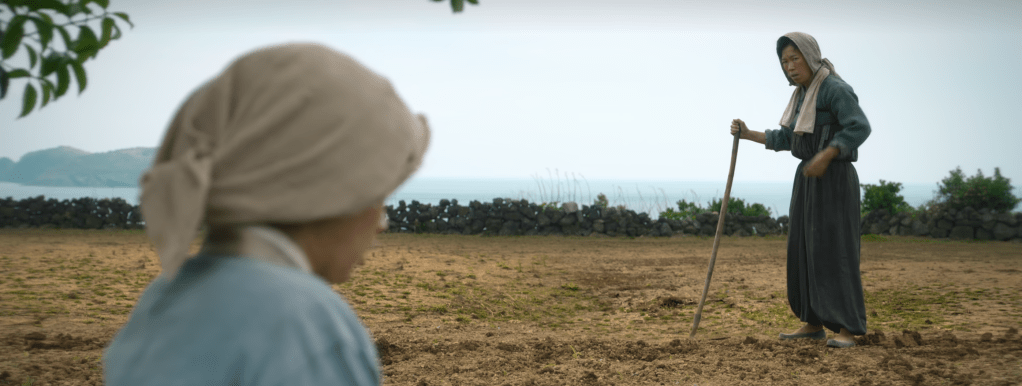 Two women working in a field on Jeju Island, with a coastal landscape visible in the background.