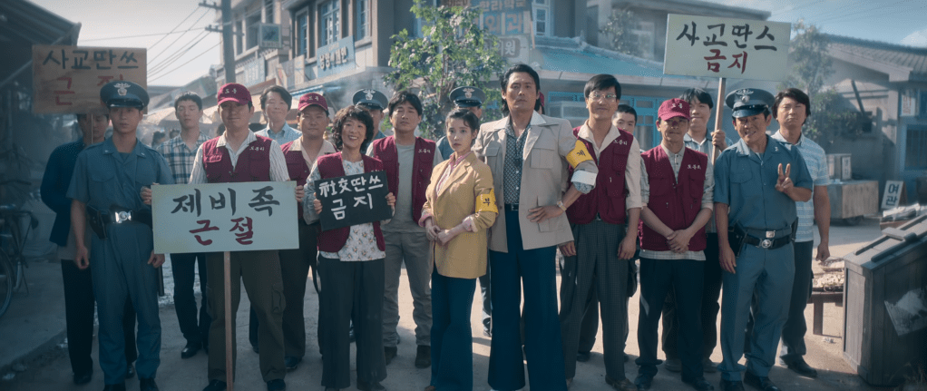A group of people, including a police officer, holding protest signs in a rural setting, showcasing community activism in a historical context.