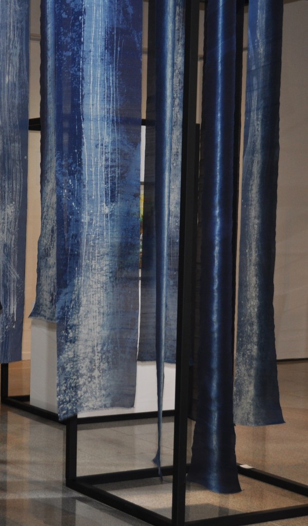 An art installation featuring vertical blue fabrics hanging from a black frame, showcasing a gradient of colors and textures.