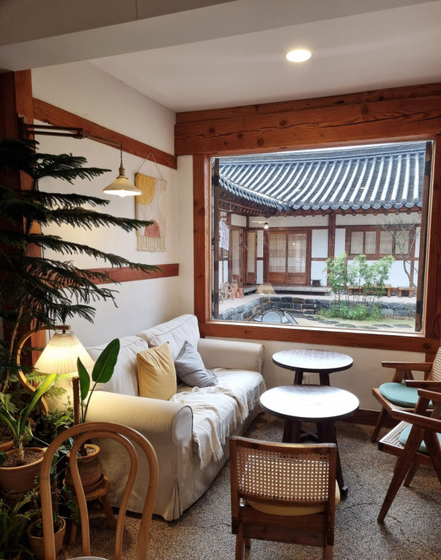 Cozy interior of a cafe featuring a white sofa, small tables, and a view of a traditional courtyard with wooden architecture and plants.