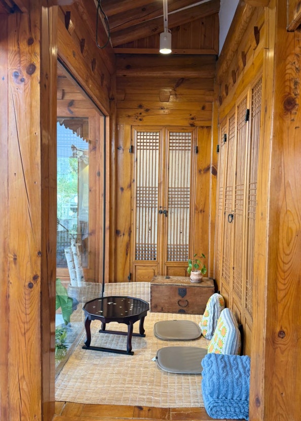Interior view of a wooden hallway with traditional Korean design, featuring sliding wooden doors, a small round table, two floor cushions, and a potted plant.