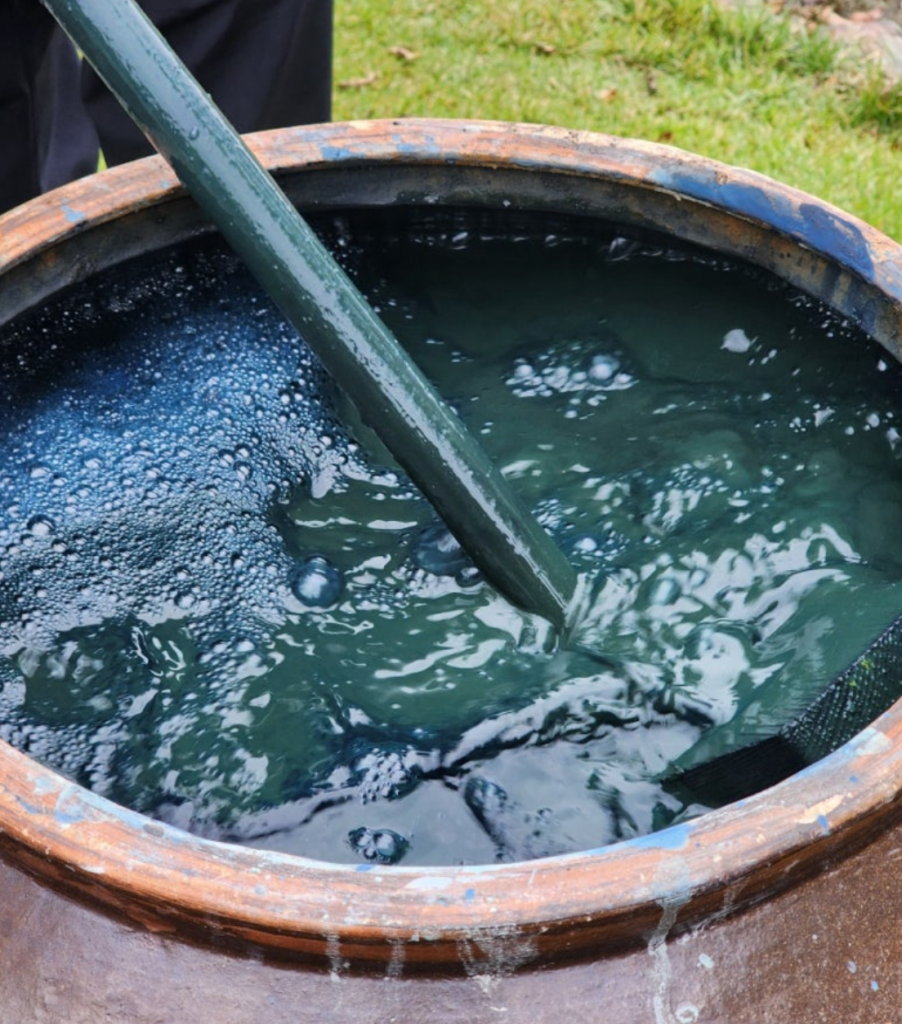 A close-up view of a large pot filled with indigo blue(jjok) liquid and bubbles, with a stirring stick submerged in the mixture.