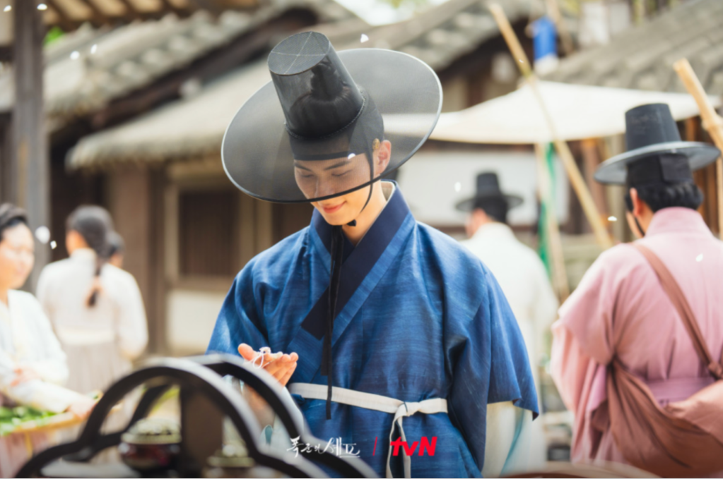 A male figure dressed in traditional Korean attire, examining an object with a focused expression, set in a historical marketplace with other figures in the background.