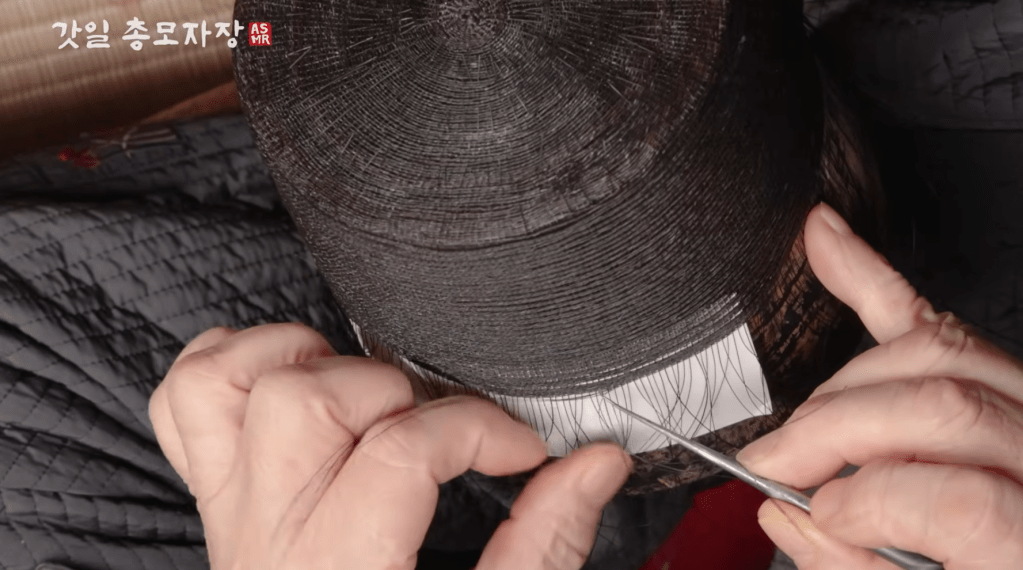 A close-up view of hands working on a traditional gat craft, using a small tool to manipulate strands of material on a circular object.