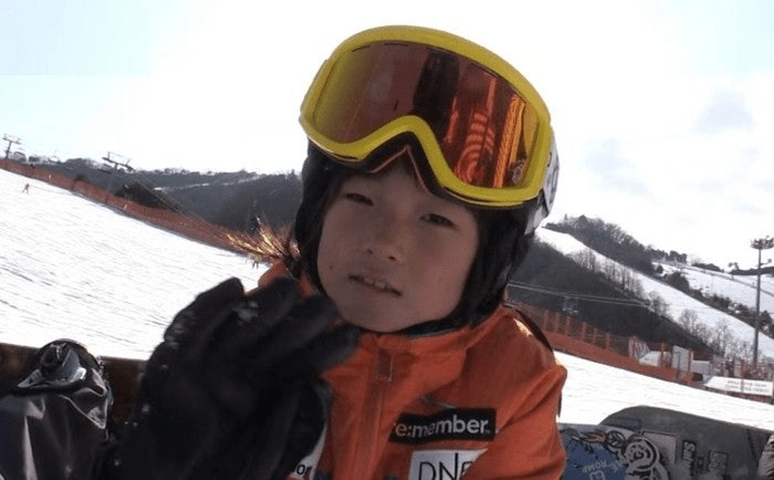 A young Choi Gaon wearing a bright orange skiing jacket and mirrored goggles, sitting on a snowboard with a snowy slope in the background.