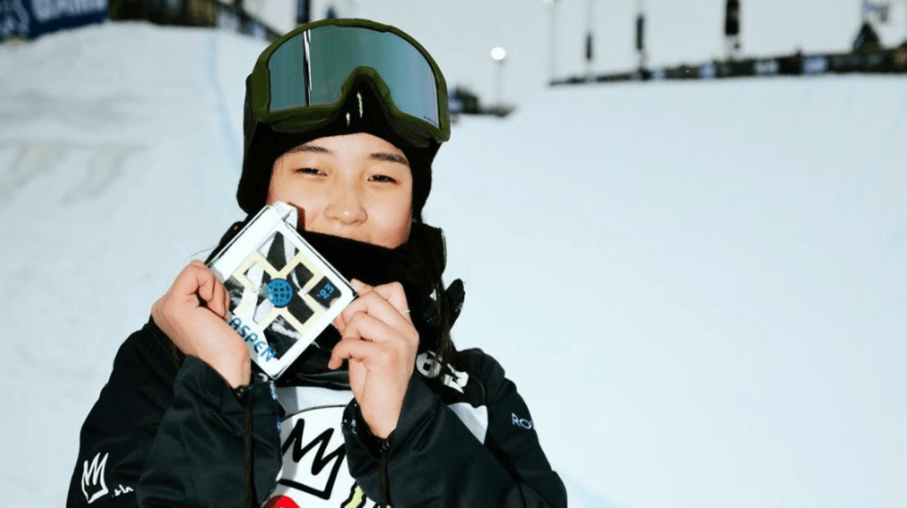 A young Choi Gain wearing winter sports gear and goggles is smiling while holding a medal in winter X Games 2023, standing in front of a snow-covered slope.