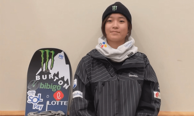 A young Choi Gaon wearing a black beanie and a striped jacket, sitting next to a snowboard adorned with various logos including Monster and Burton.