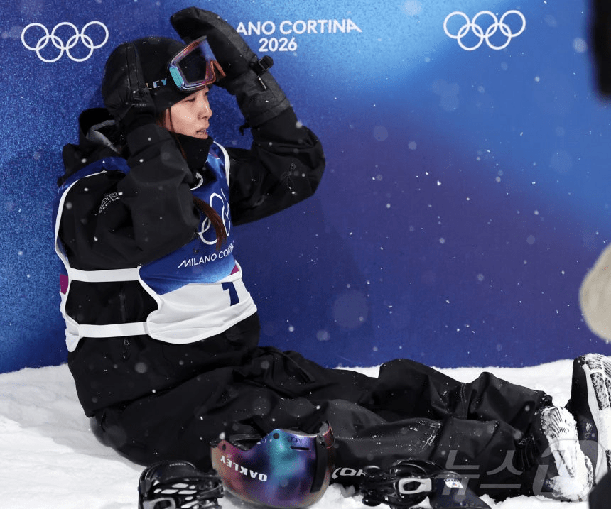 An athlete sitting on the snow, wearing snowboarding gear and goggles, with hands on their head, in front of a blue Olympic backdrop with the text 'Milano Cortina 2026'.