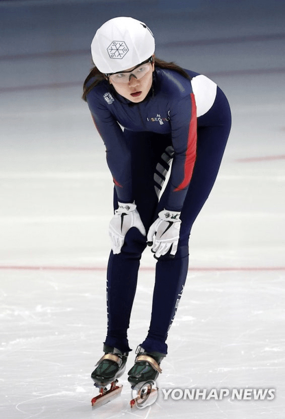 Sim Suk-hee speed skater in a blue racing suit and helmet is bending forward on the ice, focused and in a competitive stance, wearing white gloves and skate blades.