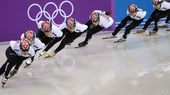 A group of speed skaters wearing matching uniforms and helmets racing in unison on an ice rink during the Olympics.