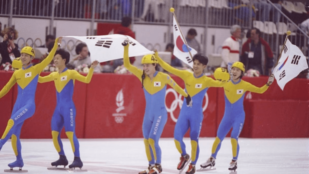 A team of Korean speed skaters celebrating on the ice, wearing colorful racing suits and holding flags of South Korea during 1992 Albertville Games.